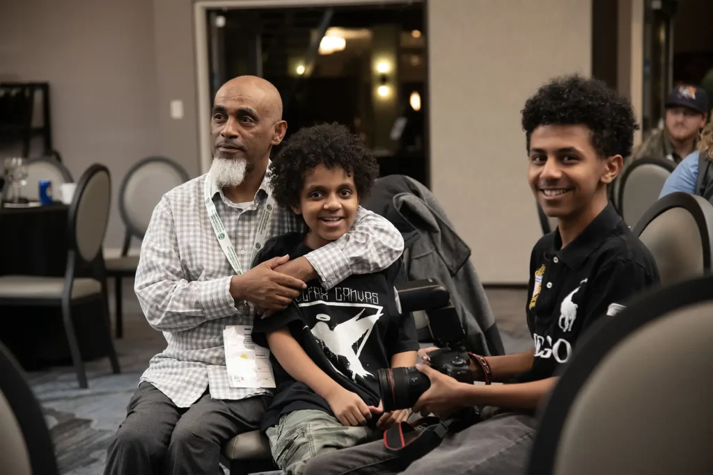 A man wraps his arm around his teenage son while the other holds a professional camera